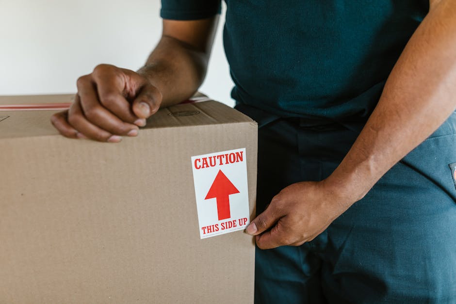 Close-up of a person wearing dark clothing during a home relocation, holding a cardboard moving box labeled with a red and white caution sticker that reads 'Caution, This Side Up' with a red arrow poi